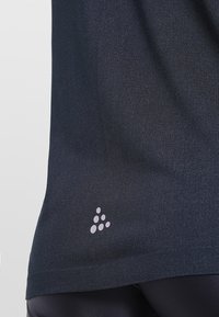 Tmavě námořnická sportovní košile vyrobená z pružné tkaniny, s jemným leskem a malým bílým logem umístěným poblíž lemu.