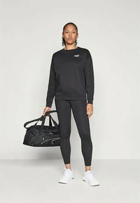 Donna in felpa nera Puma e leggings, che tiene in mano una borsa da palestra nera Puma, indossando sneakers bianche, in piedi contro uno sfondo semplice.
