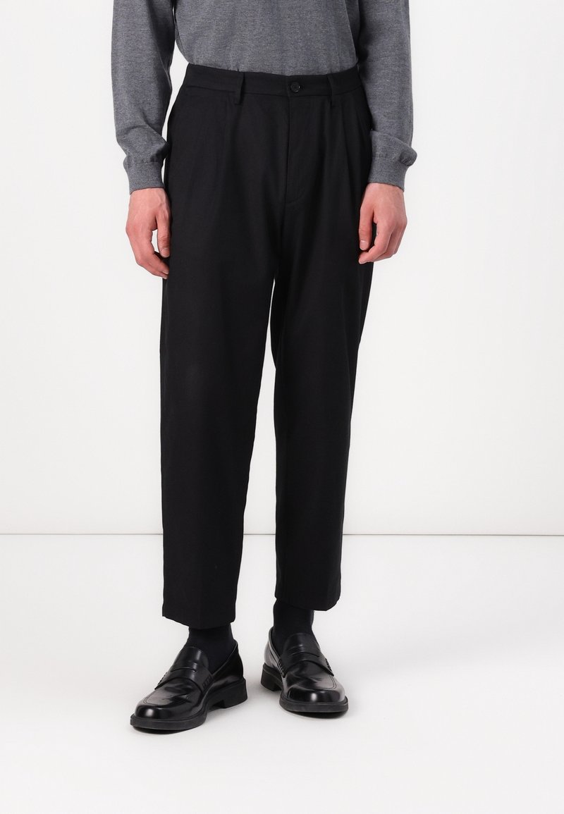 Personne portant un pantalon noir plissé, des chaussettes noires, des mocassins noirs et une chemise grise à manches longues, debout devant un fond blanc uni.