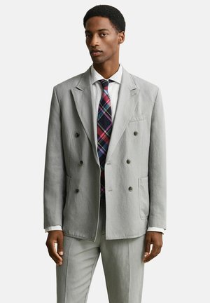 Man die een lichtgrijs dubbelbreasted pak draagt met een wit overhemd en een multicolor geruite stropdas, staand tegen een effen achtergrond.