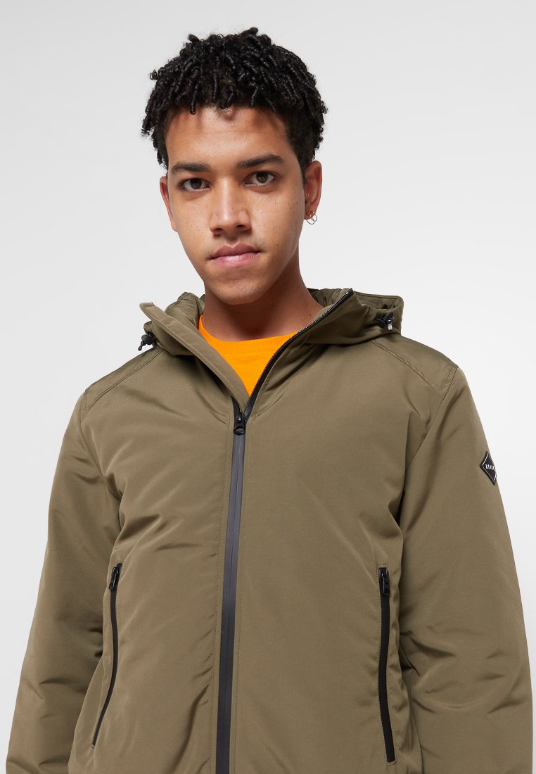 Giacca isolante verde oliva con zip frontale, cappuccio regolabile e tasche con zipper nere. Tessuto liscio con una patch del logo sulla manica.