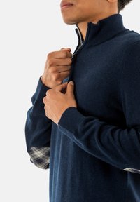 Marine ritsvest met hoge kraag, gebreide textuur en contrasterende geruite stof zichtbaar bij de manchetten. Close-up van handen die de rits aanpassen.