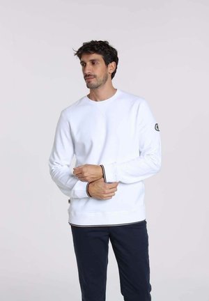 Sweatshirt blanc avec des poignets et un ourlet côtelés, présentant une petite étiquette avec logo sur la manche gauche et un col rond. Porté avec un pantalon foncé.