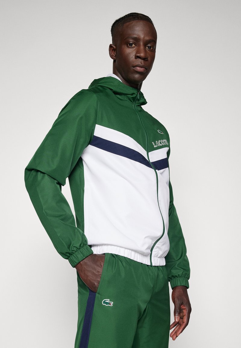 Tuta sportiva verde e bianca con strisce blu navy e logo Lacoste, composta da giacca con zip e pantaloni con vita elasticizzata.