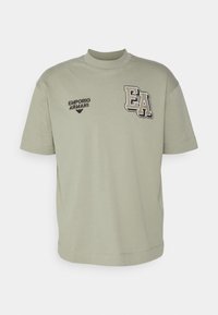 Jasnozielona bawełniana koszulka z luźnym krojem, wysokim dekoltem i haftowanymi detalami logo w czerni i bieli z przodu.