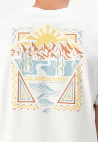 Hvit T-skjorte med et fargerikt grafisk design som inkluderer fjell, sol, kaktuser og geometriske mønstre i oransje, blått og gult. Quiksilver-logo er inkludert.