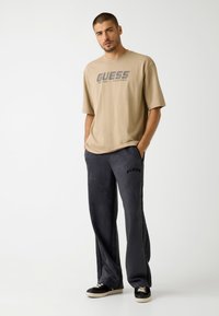 Beige cotton t-shirt with "GUESS" logo, paired with gray velour sweatpants featuring side stripes and logo detail. Black sneakers complete the look.