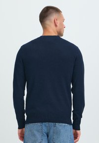 Férfi háttal áll, sötétkék, hosszú ujjú pulóverben és világoskék farmernadrágban, fehér háttér előtt.