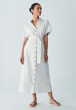 Wit-beige gestreepte, knoopbare jurk. Korte mouwen, een getailleerde taille met een bijpassende riem en voorzakken. Platte, gestrikte sandalen.