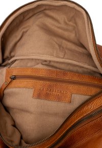 De binnenkant van een bruinleren tas met een gladde, tan kleurige stofvoering. Beschikt over een tan kleurig ritsvak en een gelabelde leren patch.