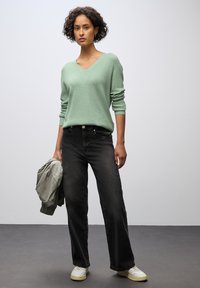 Ljusgrön v-ringad tröja i avslappnad passform. Mörka svarta jeans med rak passform. Grå bomberjacka hängande i ena handen, vita sneakers.