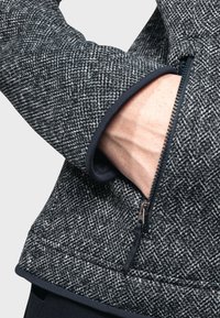 Chaqueta gris de textura con un patrón de espiga, bolsillo con cremallera y detalles en contraste en azul marino a lo largo de los puños y el dobladillo.