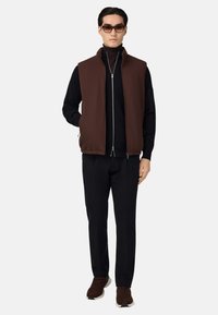 Hnedá zateplená vesta cez čierny rolák, skombinovaná s čiernymi nohavicami a hnedými teniskami; obsahuje zips a bočné vrecká.