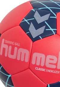 Träningsbollen har en livlig design i rött och marinblått, texturerad yta, hexagonala paneler och tryckt varumärkeslogotyp i vitt med randiga detaljer.