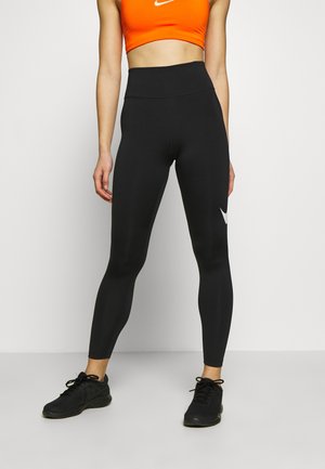 Kobieta w czarnych legginsach z wysokim stanem z białym logo Nike na nodze, w pomarańczowym staniku sportowym Nike i czarnych butach biegowych, stojąca na szarym podłożu.