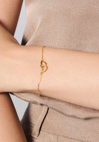 Złota bransoletka z wzorem łańcucha, z sercowym zapięciem i małymi przezroczystymi akcentami, noszona na opalonym ramieniu na neutralnym tle.