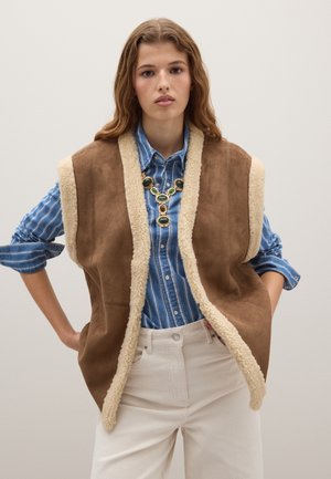 Chaleco de ante marrón con forro de piel sintética color crema, combinado con una camisa a rayas azul y blanca, y pantalones color crema.