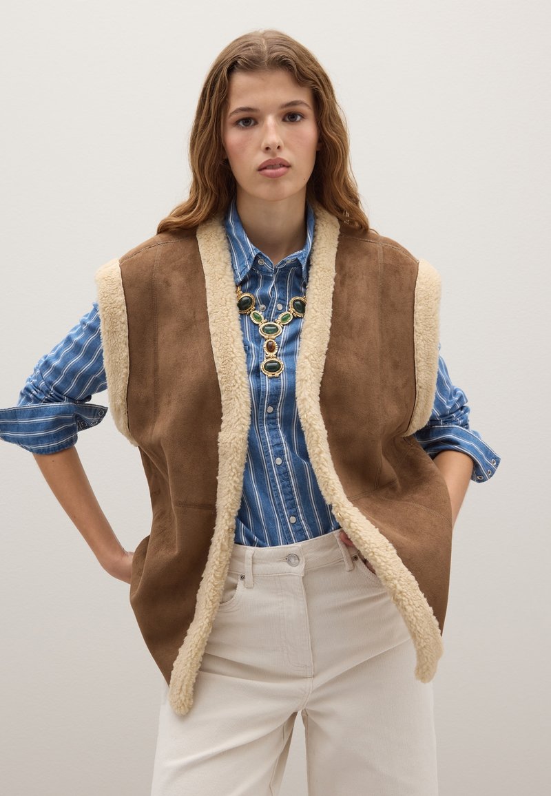 Braune Wildlederweste mit einem cremefarbenen Kunstfellfutter, über einem blau-weiß gestreiften Hemd getragen und kombiniert mit cremefarbenen Hosen.