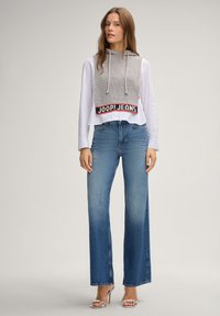 Grau gestrickter, gekürzter Hoodie mit Kordelzug und rotem Logo, über einem weißen Hemd mit Knöpfen getragen und kombiniert mit blauen Jeans mit weitem Bein.