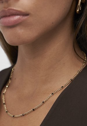 Collier en chaîne dorée avec des accents ronds noirs, des maillons plats à surface texturée, porté avec un haut brun.