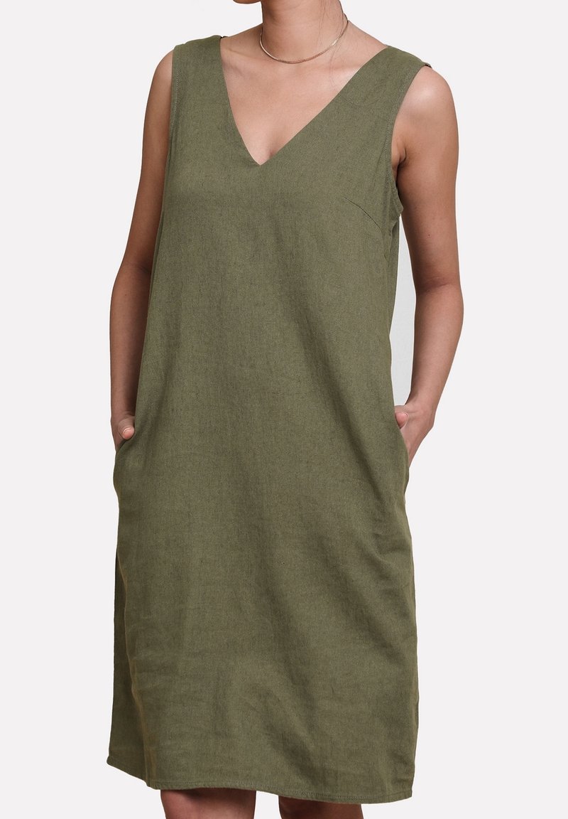 Robe sans manches vert olive avec col en V et coupe décontractée portée par une personne les mains dans les poches latérales sur fond uni.