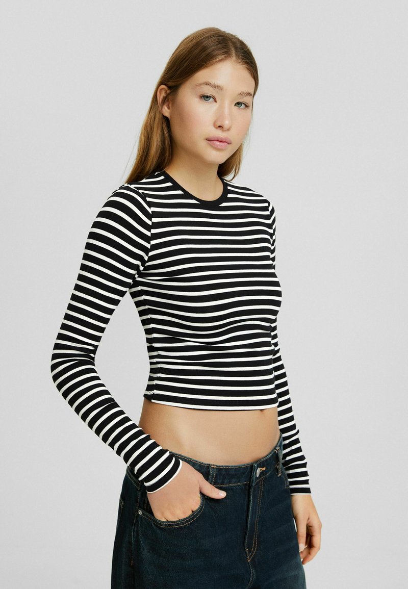 Schwarz-weißer, gestreifter Langarmoberteil mit figurbetontem Design, rundem Halsausschnitt und cropped Länge, kombiniert mit dunkelblauen Jeans.