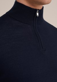 Suéter azul marino con cuello alto de cremallera, confeccionado en un tejido de punto texturizado, que presenta un corte ajustado y un diseño minimalista.