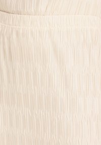 Tissu de couleur crème avec plis verticaux et taille élastique froncée, mettant en valeur la texture et les détails extensibles.
