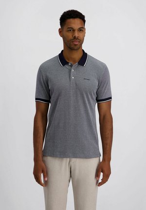 Mann mit kurzen lockigen Haaren, der ein graues Poloshirt mit schwarzem Kragen und beigen Hosen trägt, vor einem einfarbigen grauen Hintergrund steht.