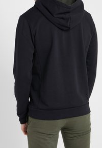 Persona con una sudadera negra y pantalones verde olivo, vista desde atrás contra un fondo sencillo.