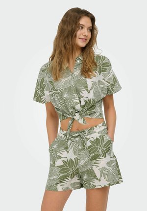 Młoda kobieta w zielono-białej, tropikalnej koszuli typu crop top zawiązanej z przodu i dołączonych szortach, stoi z rękami w kieszeniach, patrząc na bok.