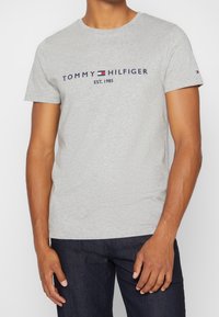 Camiseta de algodón gris con mangas cortas, que presenta el logotipo "TOMMY HILFIGER EST. 1985" en color marino, situado centralmente en la parte delantera.