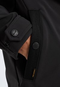 Black jacket featuring a textured fabric, buttoned cuff, and an accent patch with the text "PME Legend" in gold.