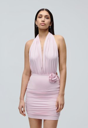 Mujer con cabello largo y oscuro que lleva un vestido sin mangas rosa pálido fruncido, con un escote en V profundo y una flor de tela en la cintura, de pie frente a un fondo gris.