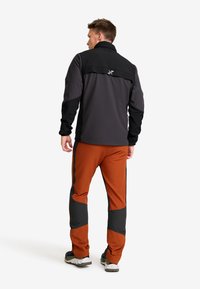 Man som vänder ryggen åt, iklädd en svart och mörkgrå jacka samt bränt orangea byxor med svarta lappar, stående framför en vit bakgrund.