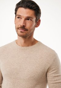 Maglione beige lavorato a maglia con scollo rotondo e polsini a costine. Presenta un logo discreto sul petto. Texture liscia e design aderente.