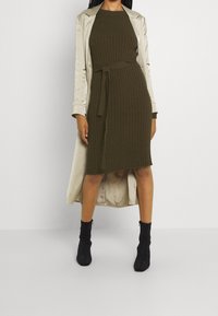 Abito a costine verde oliva con vita alta e cintura, abbinato a un trench coat beige chiaro. La modella indossa stivali neri alla caviglia.