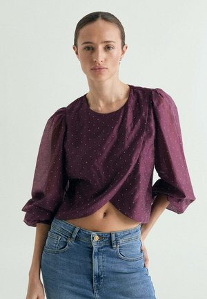 Mujer con el pelo recogido que lleva una blusa morada con mangas abullonadas de lunares y vaqueros azules sobre un fondo claro y liso.