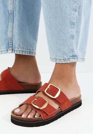 Voeten met oranje suède sandalen met twee grote gouden gespen, gecombineerd met lichtblauwe cropped spijkerbroeken.