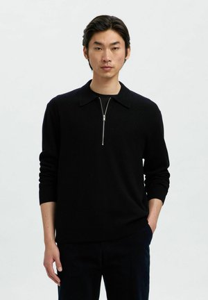 Pull en maille noir avec un col zippé, des manches longues et une coupe décontractée. Le tissu semble lisse avec une texture douce.