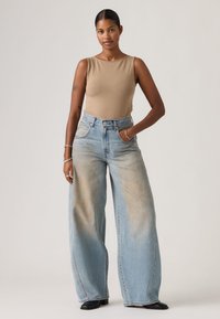 Γυναίκα που στέκεται με το δεξί χέρι στην  τσέπη, φορώντας μπεζ αμάνικο τοπ, φαρδιά ξεθωριασμένα μπλε τζιν παντελόνια και μαύρα παπούτσια σε ανοιχτό φόντο.