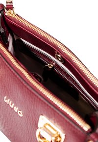 Borsa in pelle bordeaux con hardware dorato, chiusura con zip e interno foderato. Presenta un logo impresso all'esterno.