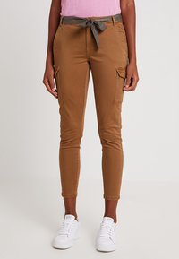 Bruna cargobyxor med en figurnära design, framfickor och ett knytet grått bälte. Bärs med vita sneakers mot en neutral bakgrund.