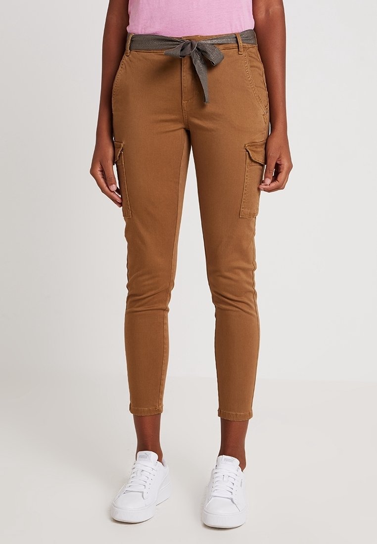 Bruna cargobyxor med en figurnära design, framfickor och ett knytet grått bälte. Bärs med vita sneakers mot en neutral bakgrund.