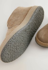 Chaussures en daim marron avec un bout rond, dotées d'une semelle en caoutchouc gris texturé et de coutures visibles le long du bord. Deux chaussures sont présentées.