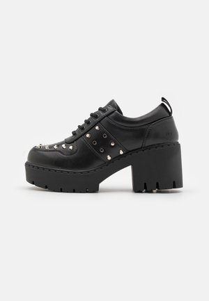 Zwarte leren plateau schoen met dikke chunky hak, veters aan de voorkant en zilverkleurige metalen studs aan de zijkanten en teengebied, zijaanzicht op witte achtergrond.