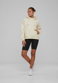 Felpa crema con tasca frontale e cordino regolabile. Gli shorts neri da bici e le sneakers bianche completano l'outfit. Texture liscia.