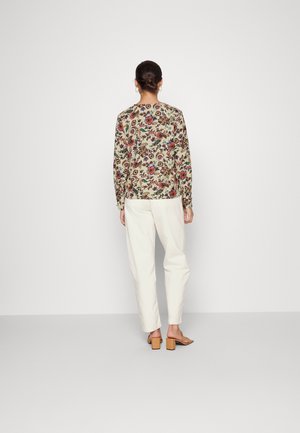 Femme avec les cheveux attachés en chignon, portant un haut à manches longues à fleurs, un pantalon blanc taille haute et des sandales à talons beige, debout de dos.