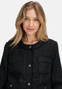 Kobieta z blond włosami, ubrana w czarną teksturowaną kurtkę zapinaną na guziki, na jednolitym jasnym tle, patrzy na wprost i delikatnie się uśmiecha.