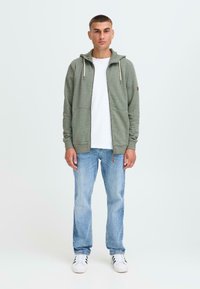 Zelena zip-up hoodie s vezicama, nosi se preko bijele majice. Uparen s svijetloplavim trapericama i bijelim tenisicama. Glatka, ležerna tkanina.